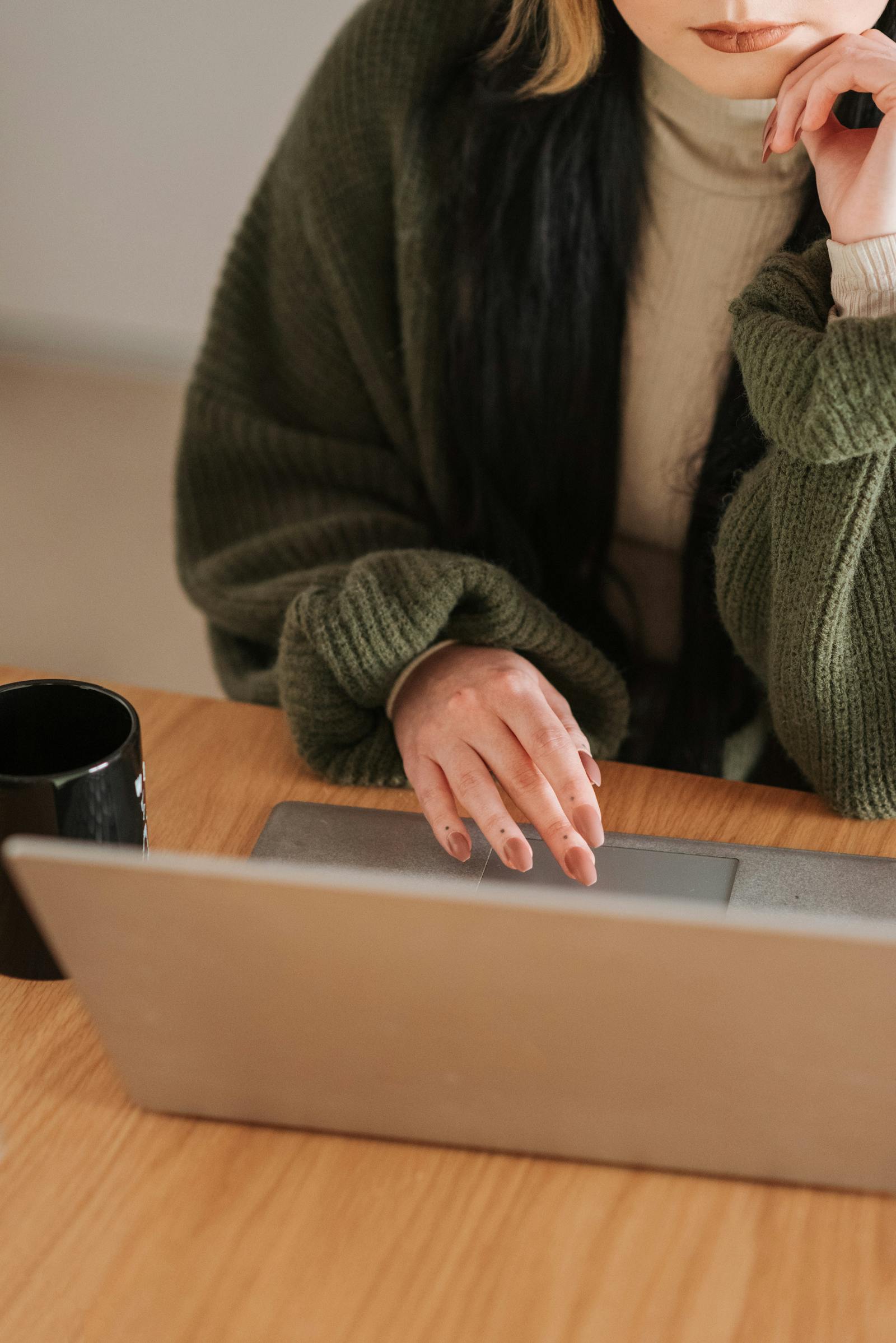 A person in a green knit cardigan rests her hand at her chin, the other on a laptop trackpad, working at a wooden table with a coffee mug