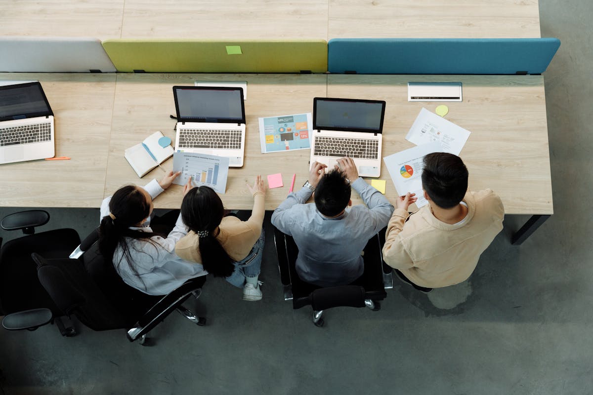 Overhead view of four colleagues collaborating on laptops and printed charts at a long shared desk with sage and blue panels